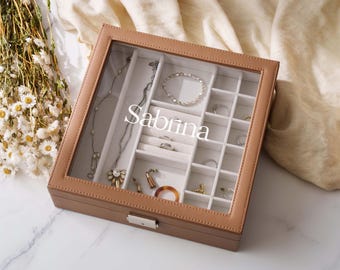 Puede incluir: Un joyero cuadrado de color tostado con una tapa de cristal transparente que presenta el texto blanco Sabrina. El interior está forrado con tela blanca y dividido en múltiples compartimentos que contienen varios collares, anillos y pulseras.