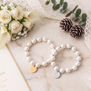 a bouquet of white roses, two pearl bracelets, pine cones, and a wedding invitation on a marble surface.