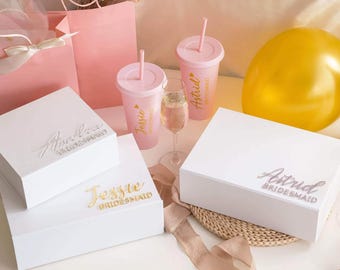 May include: Three white rectangular gift boxes with personalised names in gold or silver script, two light pink tumblers with straws labelled Jessie and Astrid, a clear glass of champagne, a pink gift bag, and a yellow balloon.