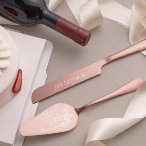 a pink cake knife and a pink cake server with the words "Let's Eat Cake" engraved on them, along with a bottle of wine and a white ribbon.