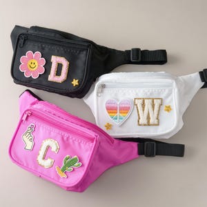 Pode incluir: Três pochetes em preto, branco e rosa com vários patches e letras. A pochete preta tem um rosto sorridente amarelo, um D rosa e uma estrela dourada. A pochete branca tem um coração com um sinal de paz arco-íris, um W dourado e duas estrelas douradas. A pochete rosa tem um gesto de mão rosa, um C dourado e um cacto verde.