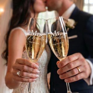 Può includere: Due flûte da champagne in vetro trasparente riempite di liquido frizzante. Il bicchiere sinistro è inciso con Mrs. Melville e July 25, 2026. Il bicchiere destro è inciso con Mr. Melville e July 25, 2026. Entrambi i bicchieri hanno stelo sottile.