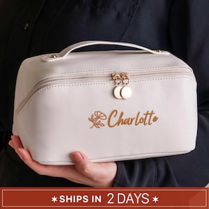 May include: A cream-colored cosmetic bag with a top handle and a zipper closure. The bag features gold-colored hardware and the name "Charlotte" embroidered in gold script. The bag is held in a person's hands.