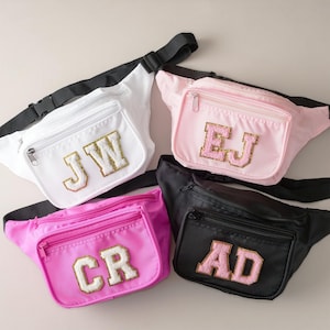 Op de afbeelding: Vier heuptasjes in verschillende kleuren met gepersonaliseerde initialen in gouden glitterletters. De heuptasjes zijn wit, roze, felroze en zwart.