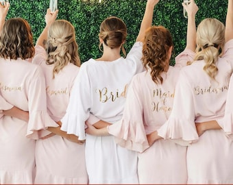 May include: Five individuals stand in a row wearing matching robes with ruffled sleeves and hems. The center robe is white with the gold text Bride, while the surrounding four robes are light pink with gold text reading Bridesmaid or Maid of Honor.