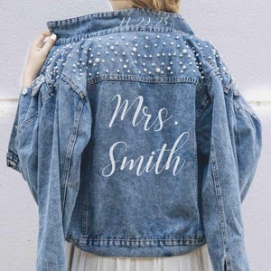 May include: A blue denim jacket featuring white pearl embellishments across the upper back and shoulders. The back of the jacket displays the white text "Mrs. Smith" in a cursive script, with the date "11.20.23" printed along the collar seam.