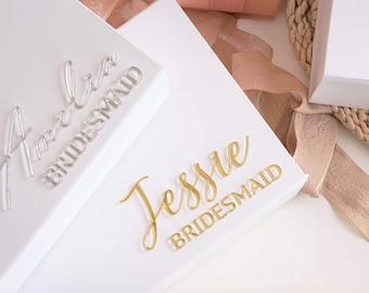 May include: Two white rectangular gift boxes featuring raised lettering. One box displays the name Amelia in silver script with the word BRIDESMAID in block letters. The other box features the name Jessie in gold script with BRIDESMAID below.