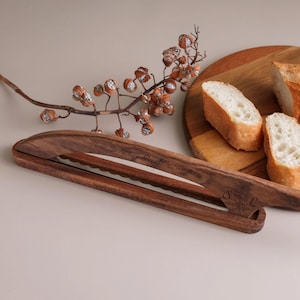 Könnte beinhalten: Ein dunkelbraunes Brotmesser aus Holz mit geschwungenem Griff und gezahnter Klinge. Der Griff ist mit dem Text "Home Is Where Bread Is Warm" und "Baked with Love, Just for Victoria" sowie den Initialen "S L" graviert.