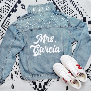 Puede incluir: Una chaqueta de mezclilla azul claro con perlas blancas en la parte superior de la espalda y los hombros. El cuello muestra la fecha 11. 20. 23 en texto blanco, mientras que el panel trasero presenta el texto blanco Mrs. Garcia. Zapatillas blancas.