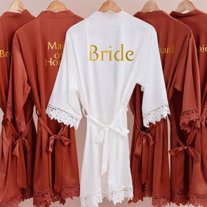May include: A selection of satin robes, including a white robe with "Bride" in gold lettering and several rust-coloured robes with gold lettering. The white robe has lace trim on the sleeves and a matching tie. The rust-coloured robes have lace trim and matching ties.
