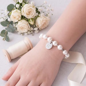 A bracelet with white pearls and a charm is worn on a person's wrist, accompanied by a bouquet of white roses, a spool of thread, and a gift box with a white ribbon.