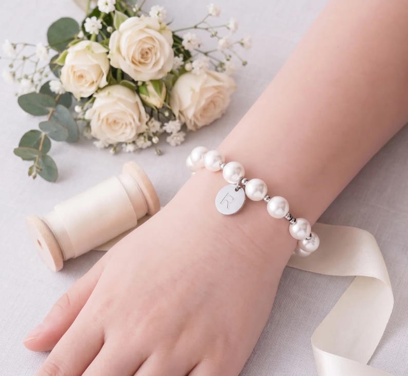 A bracelet with white pearls and a charm is worn on a person's wrist, accompanied by a bouquet of white roses, a spool of thread, and a gift box with a white ribbon.