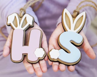 Könnte beinhalten: Zwei hölzerne Buchstaben-Anhänger, ein H in Pastellrosa und ein S in Pastellblau. Beide verfügen über weiße Hasenohren an der Oberseite, einen kleinen weißen Schwanz an der Seite und eine Schlaufe aus Juteschnur zum Aufhängen aus Schichtholz.