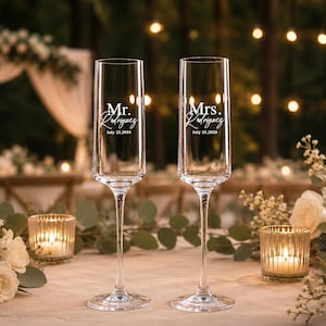 May include: Two clear glass champagne flutes with slender stems and bases. The left glass features white text reading Mr. Rodriguez July 25, 2026, and the right glass features white text reading Mrs. Rodriguez July 25, 2026, in a mix of serif and script fonts.