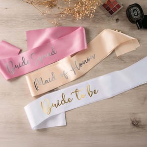 May include: Three satin sashes in pink, beige, and white, with metallic lettering. The pink sash reads "Bride Squad", the beige sash says "Maid of Honor", and the white sash says "Bride to be".