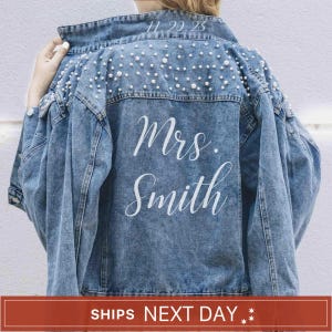 May include: A blue denim jacket featuring white pearl embellishments across the upper back and shoulders. The back of the jacket displays the white printed text "Mrs. Smith" in a script font, with the date "11.20.23" printed above the text.