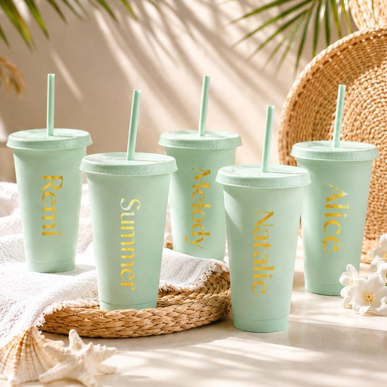A group of mint green reusable cups with personalized names on them, placed on a woven basket and surrounded by white flowers and a straw hat.