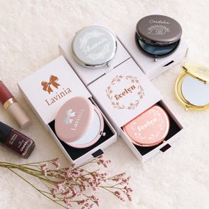 May include: A collection of round compact mirrors in silver, gold, rose gold, and matte pink finishes, each personalized with names like Lavinia, Evelyn, Rhiannon, and Cordolia. The mirrors are presented in white sliding gift boxes with matching prints.