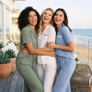 May include: Three women wearing matching pajama sets, embracing and laughing on a deck overlooking the ocean. The pajamas are in shades of green, white, and blue, with personalized names embroidered on the shirts.