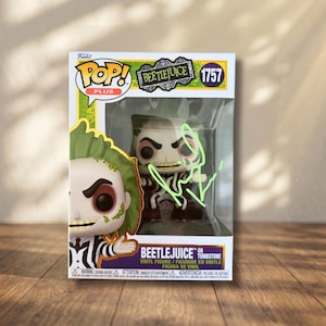 May include: A Funko Pop Plus Beetlejuice on Tombstone vinyl figure in its original packaging. The box features the character name, number 1757, and a green autograph on the clear plastic window. The figure inside wears a black and white suit.