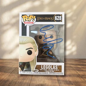 Könnte beinhalten: Eine Funko Pop Movies The Lord of the Rings Legolas Vinylfigur, Nummer 628, in ihrer Originalverpackung. Das durchsichtige Kunststofffenster ist auf der Vorderseite mit einem Autogramm in blauer Tinte versehen. Die Box zeigt den Namen.