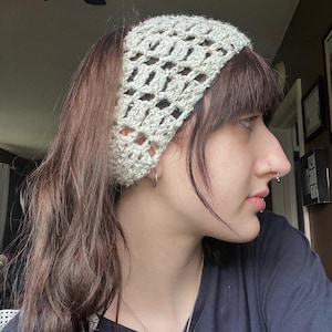 May include: A light green crocheted headband with an open weave design. The headband is wide and covers the forehead. The hair is brown and parted to the side. The person is wearing a dark blue shirt and a silver necklace.