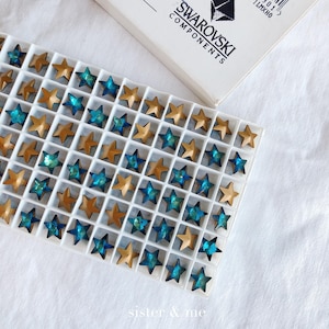 Puede incluir: Una bandeja blanca llena de piedras de cristal en forma de estrella doradas y turquesa. La bandeja está etiquetada como "Swarovski Components".