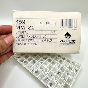 May include: A white cardboard box labeled 4861 MM 8.0 CRYSTAL COMET ARG.LIGHT VZ 2/00/00 GROSS = 288 PCS. SWAROVSKI COMPONENTS Made in Austria, positioned above a white plastic tray containing numerous small, clear, faceted round crystal components.