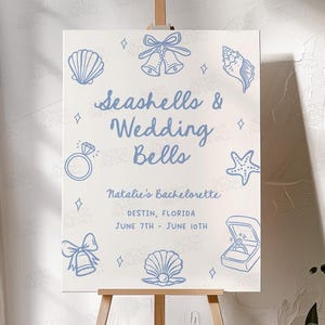 Puede incluir: Un cartel blanco con ilustraciones azules de conchas marinas, campanas de boda y anillos. El texto dice "Seashells & Wedding Bells" y "Natalie's Bachelorette, Destin, Florida, 7 de junio - 10 de junio".