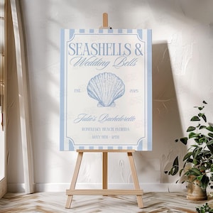 Op de afbeelding: Een blauw en wit gestreept bord met de tekst "Seashells & Wedding Bells" en een schelpillustratie. Het bord bevat ook de tekst "Julie's Bachelorette", "Rosemary Beach, Florida", en "May 9th - 12th".