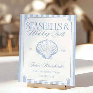 Op de afbeelding: Een lichtblauw en wit bord met de tekst "Seashells & Wedding Bells" en een schelpillustratie. Het bord bevat ook de tekst "Julie's Bachelorette", "Rosemary Beach Florida" en "May 9th - 12th".