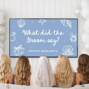 May include: A light blue sign with the text "What did the Groom say?" and "Natalie's Bachelorette" in white script. The sign is decorated with seashell and ring illustrations. Five women are seated in front of the sign.