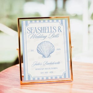 Op de afbeelding: Een lichtblauw en wit gestreept bord met de tekst "Seashells & Wedding Bells" in een decoratief lettertype. Het bord bevat ook een schelpillustratie, de tekst "Julie's Bachelorette", "Rosemary Beach Florida" en "May 9th - 12th".