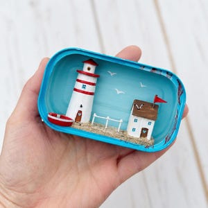 May include: Miniature coastal scene in a blue container. A white and red striped lighthouse stands next to a small house with a red flag. A red boat sits near the lighthouse, and seagulls fly in the blue sky. The scene is set on a sandy beach.