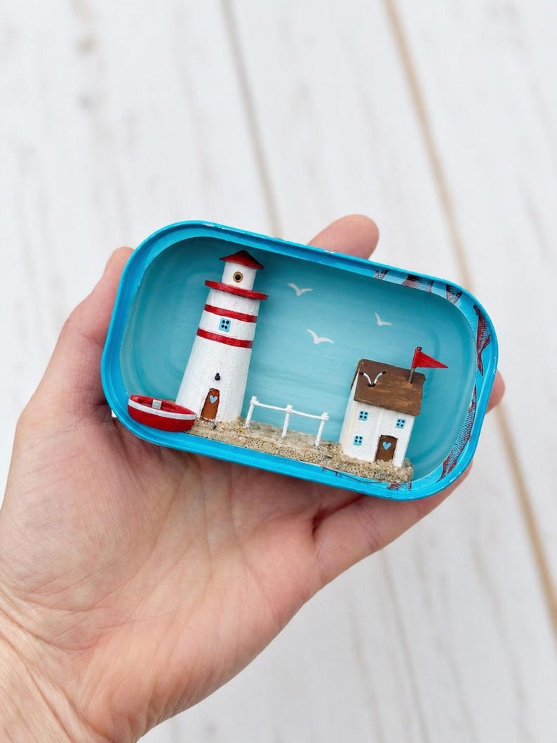 May include: Miniature coastal scene in a blue container. A white and red striped lighthouse stands next to a small house with a red flag. A red boat sits near the lighthouse, and seagulls fly in the blue sky. The scene is set on a sandy beach.