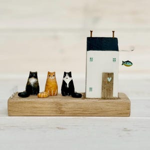 May include: A decorative wooden block display featuring three small cat figurines in black, orange, and black-and-white colors, positioned next to a miniature white house with a black roof, a wooden door, and a small hanging blue fish ornament.