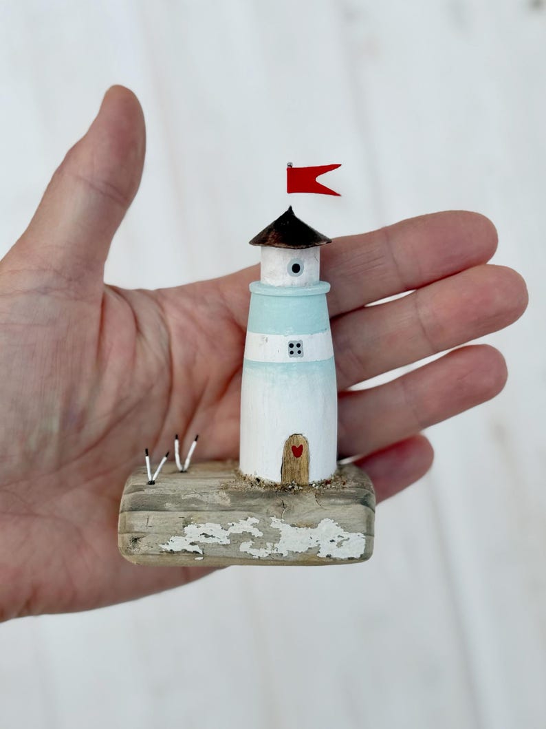 Pu&ograve; includere: Una statuina a forma di faro in miniatura, dipinta di bianco e azzurro chiaro, con una bandiera rossa in cima. Il faro ha una piccola porta di legno con un cuore e poggia su una base di legno invecchiato. Due piccoli accenti bianchi e neri sono sulla base.