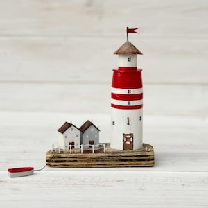 Op de afbeelding: Een decoratief houten vuurtorenbeeldje met rode en witte horizontale strepen, een kleine rode vlag op de top en een reddingsboei op de deur. Twee kleine grijze huisjes staan naast de vuurtoren op een gelaagde houten basis met een rood bootje.