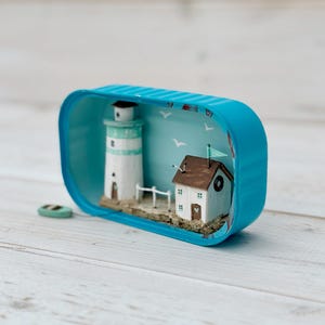 May include: A miniature lighthouse and house scene inside a blue tin can. The lighthouse is white with a green roof. The house is white with a brown roof and a small window. The scene is set against a blue background with white birds and a blue sky.