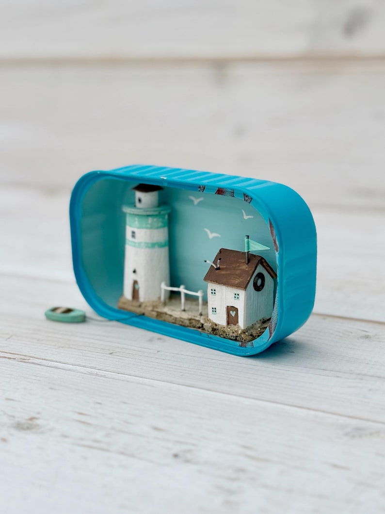 May include: A miniature lighthouse and house scene inside a blue tin can. The lighthouse is white with a green roof. The house is white with a brown roof and a small window. The scene is set against a blue background with white birds and a blue sky.