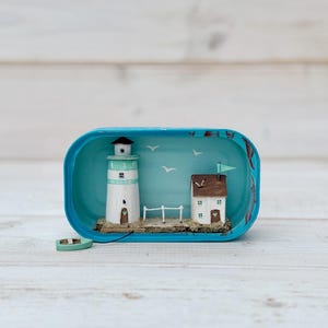 May include: A miniature lighthouse and house scene inside a blue tin can. The lighthouse is white with a green stripe and a brown roof. The house is white with brown trim and a brown roof. The scene is set on a brown base with a green boat and a white picket fence. The background is blue with white birds.
