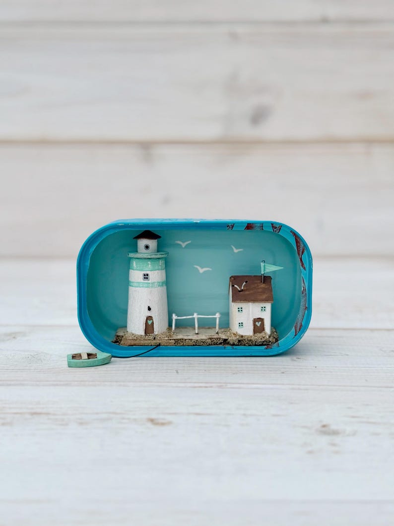 May include: A miniature lighthouse and house scene inside a blue tin can. The lighthouse is white with a green stripe and a brown roof. The house is white with brown trim and a brown roof. The scene is set on a brown base with a green boat and a white picket fence. The background is blue with white birds.