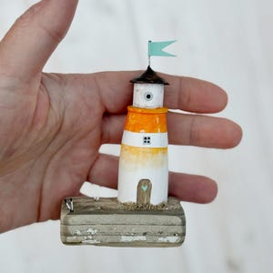 Pu&ograve; includere: Una statuetta di faro in miniatura dipinta a mano. Il faro presenta un design bianco, arancione e nero, sormontato da una piccola bandiera blu. &Egrave; montato su una base di legno invecchiato. Il faro &egrave; alto circa 7 cm.