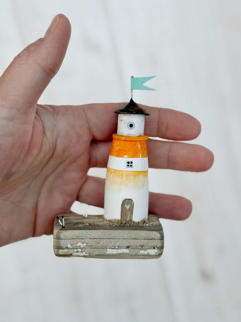 Pu&ograve; includere: Una statuetta di faro in miniatura dipinta a mano. Il faro presenta un design bianco, arancione e nero, sormontato da una piccola bandiera blu. &Egrave; montato su una base di legno invecchiato. Il faro &egrave; alto circa 7 cm.