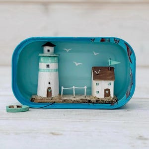May include: A miniature lighthouse and house scene inside a blue oval tin. The lighthouse is white with a light blue stripe and a brown door. The house is brown with white trim and a green roof. The scene is set against a light blue background with white birds and a fence.