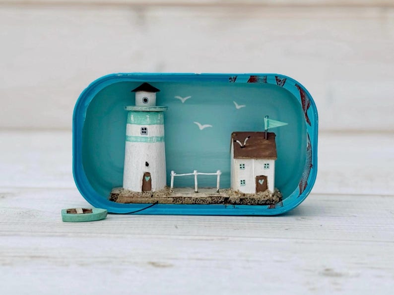 May include: A miniature lighthouse and house scene inside a blue oval tin. The lighthouse is white with a light blue stripe and a brown door. The house is brown with white trim and a green roof. The scene is set against a light blue background with white birds and a fence.