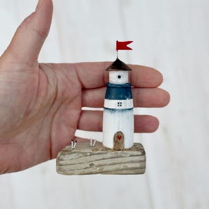 Pu&ograve; includere: Una statuetta di faro in miniatura con una bandiera rossa, un tetto marrone scuro e una fascia blu. Il faro &egrave; bianco con una piccola porta di legno e un cuore rosso. &Egrave; montato su una base di legno.