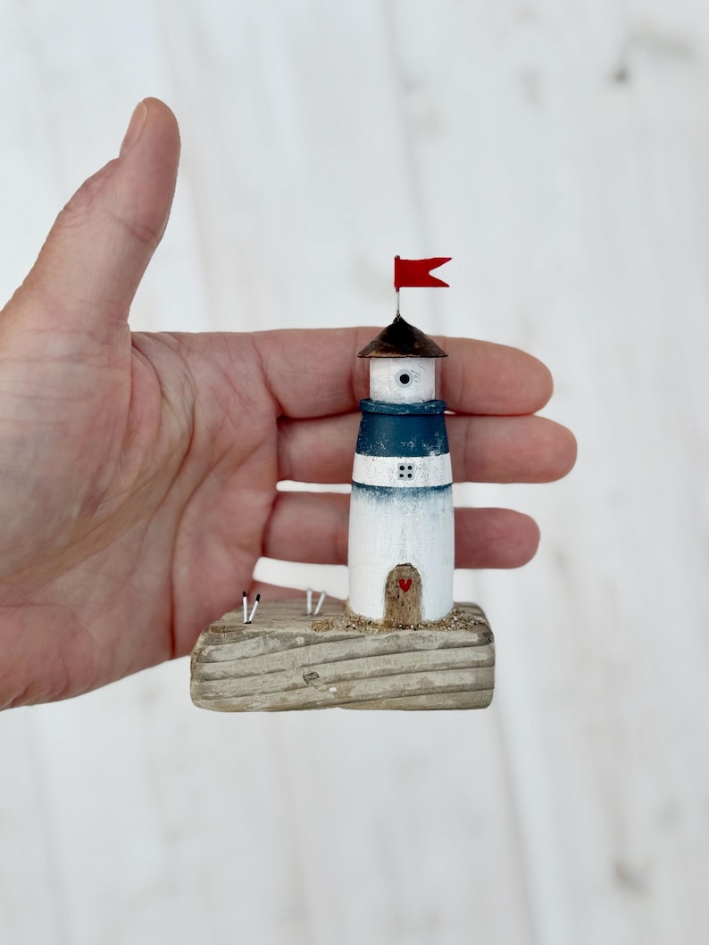 Pu&ograve; includere: Una statuetta di faro in miniatura con una bandiera rossa, un tetto marrone scuro e una fascia blu. Il faro &egrave; bianco con una piccola porta di legno e un cuore rosso. &Egrave; montato su una base di legno.
