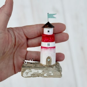 Pu&ograve; includere: Una statuetta di un faro in miniatura con una torre a strisce rosse e bianche, un tetto marrone e una piccola bandiera blu. Il faro &egrave; posizionato su una base strutturata e usurata. Una piccola porta a forma di cuore &egrave; visibile.