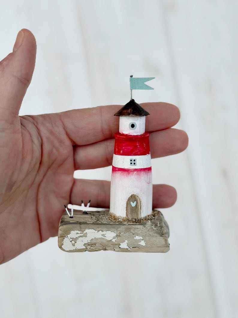 Pu&ograve; includere: Una statuetta di un faro in miniatura con una torre a strisce rosse e bianche, un tetto marrone e una piccola bandiera blu. Il faro &egrave; posizionato su una base strutturata e usurata. Una piccola porta a forma di cuore &egrave; visibile.
